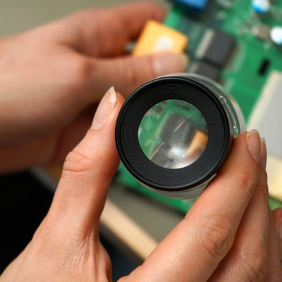PCB-Board-Cybersecurity Close-up of a magnifying glass over a PCB board.