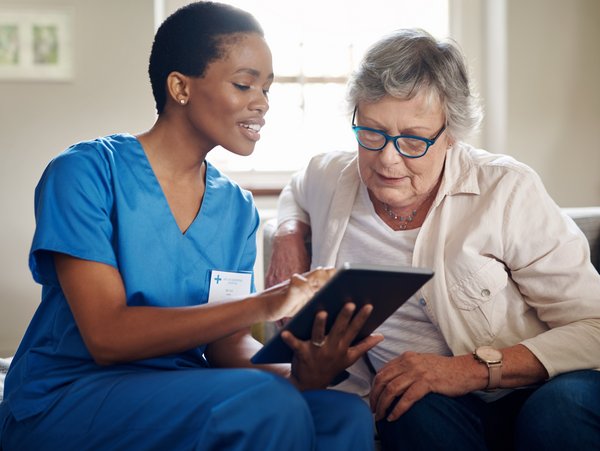 Eine medizinische Fachkraft und eine Patientin sitzen auf einer Couch und schauen auf ein Tablet.