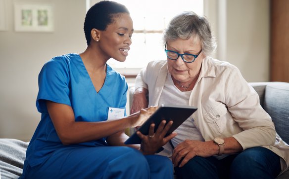 Eine medizinische Fachkraft und eine Patientin sitzen auf einer Couch und schauen auf ein Tablet.