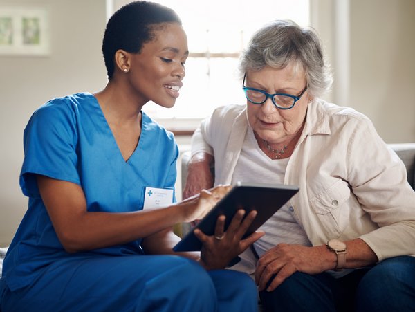 Eine medizinische Fachkraft und eine Patientin sitzen auf einer Couch und schauen auf ein Tablet.