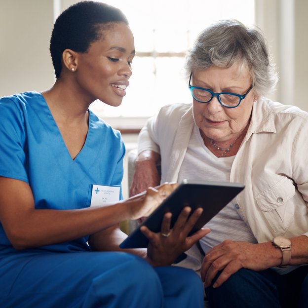 Eine medizinische Fachkraft und eine Patientin sitzen auf einer Couch und schauen auf ein Tablet.