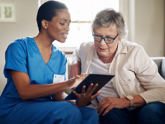 Eine medizinische Fachkraft und eine Patientin sitzen auf einer Couch und schauen auf ein Tablet.