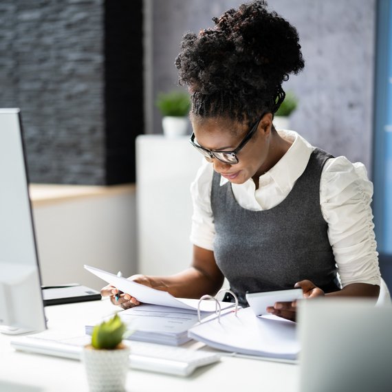 Eine Frau sitzt am Schreitische uns arbeitet mit einem Aktenorder
