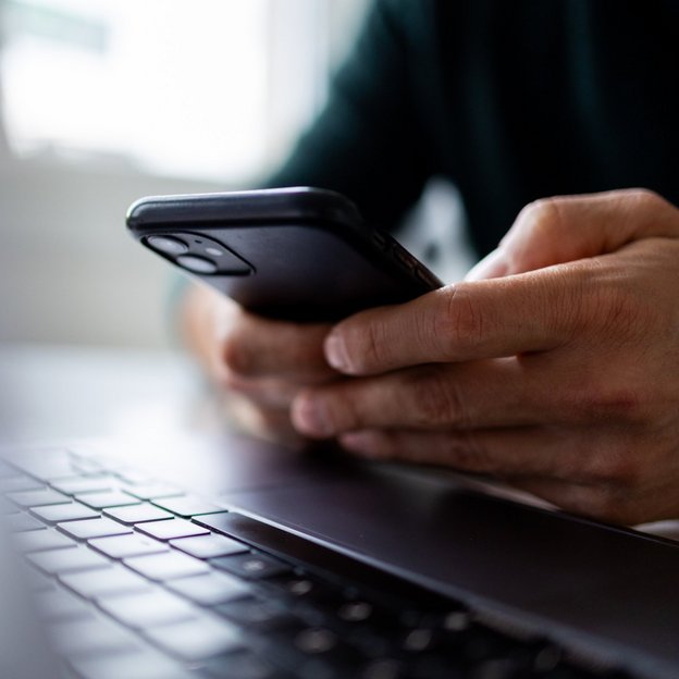 Ein Mann sitzt am Laptop und bedient sein Handy. | TÜVIT