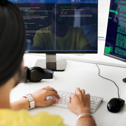 Eine Person sitzt am Schreibtisch und tippt am PC, auf den Bildschirmen sieht man Code.
