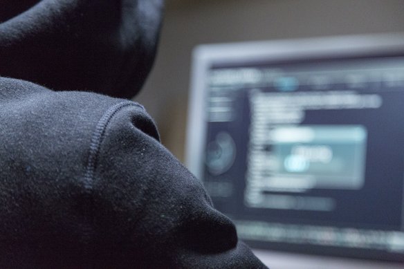 Eine Person mit Kapuzenpulli sitzt an einem Monitor mit Code.