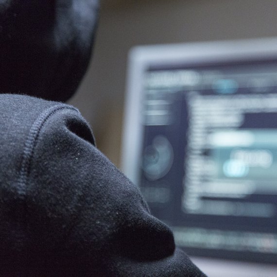 Eine Person mit Kapuzenpulli sitzt an einem Monitor mit Code.