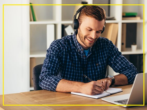 A man with headphones is sitting at the table, looking at his laptop and writing something in a notebook.