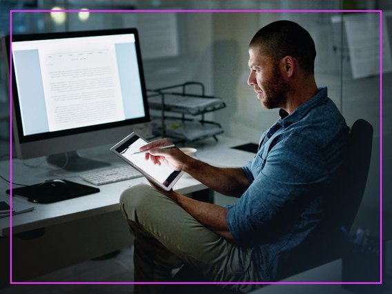 büro-tablet-pc-dokument Ein Mitarbeitender sitzt abends am Schreibtisch und arbeitet gleichzeitig am PC und am Tablet.