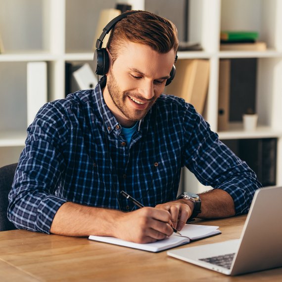 Ein Mann mit Kopfhörern sitzt am Tisch vor dem Laptop und schreibt etwas in ein Notizbuch.