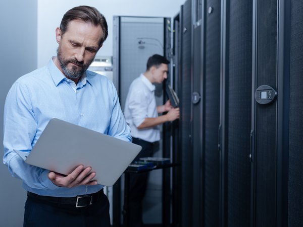Zwei Männer stehen in einem Rechenzentrum, einer tippt etwas in den Laptop, den er hält.