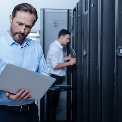 Zwei Männer stehen in einem Rechenzentrum, einer tippt etwas in den Laptop, den er hält.