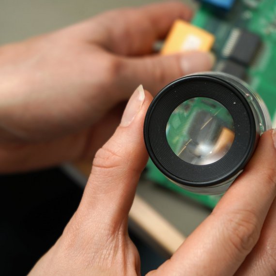 Eine Person schaut sich eine Hardwarekomponente mit der Lupe an - Hardware Labor von TÜV Informationstechnik