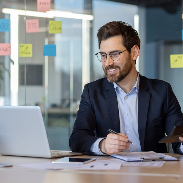 Ein Mann sitzt in einem Büro an einem Laptop und lächelt. | TÜVIT